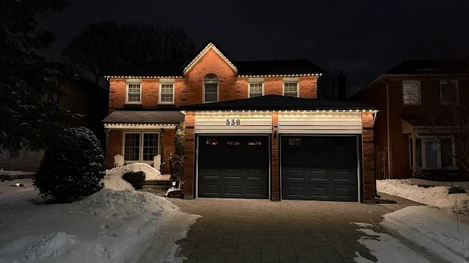 Two-storey GTA brick home with warm white permanent Christmas lights installed along the roofline and garage soffit at night.
