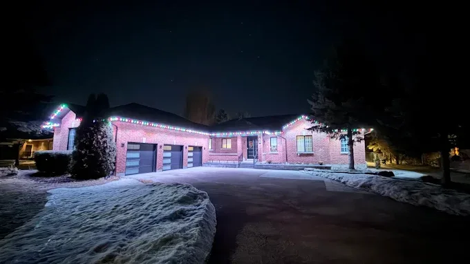 Brick bungalow with multicolour permanent Christmas lights along the roofline, lighting up the driveway and garage at night.
