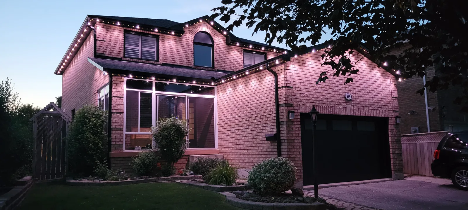 Two-storey brick GTA home with warm pink and white permanent outdoor LED roofline lighting by Toronto Glow, photographed at dusk from the front yard